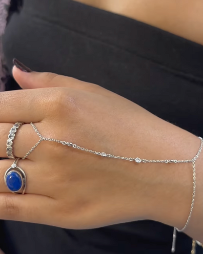 A close-up image of a hand wearing the Diamond Bezel Hand Chain in a silver finish. The delicate chain features distanced cubic zirconia stones set in bezel settings, gracefully draping across the hand. The chain wraps around the wrist and connects to a finger ring, adding a touch of sparkle against the skin, set against a black fabric background.