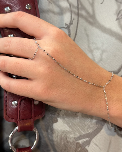 Close-up image of a hand wearing the Feather Hand Chain, a delicate piece of jewelry made from 925 sterling silver with rhodium plating. The chain wraps around the wrist and connects to a ring on the finger, showcasing its elegant and lightweight design against the skin. The hand holds a burgundy leather strap with silver studs, highlighting the contrast between the soft, shimmering chain and the textured leather.