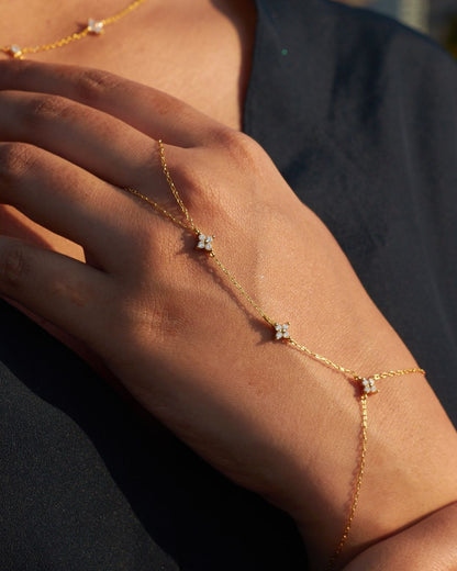 A close-up of a model’s hand wearing the In Bloom Hand Chain, featuring small cubic zirconia floral accents along a fine gold-plated chain. The sunlight catches the sparkle of the stones, adding a soft glow to the jewelry. The model's black outfit provides contrast, making the gold and crystals stand out.