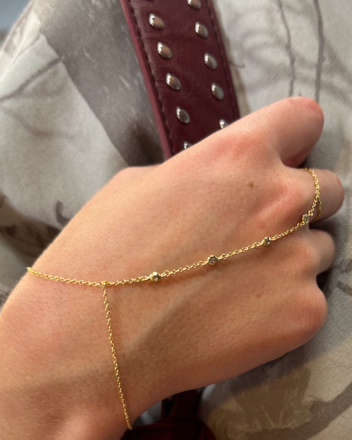 Gold Diamond Bezel Hand Chain: A close-up image of a hand wearing the Diamond Bezel Hand Chain in a gold finish. The chain features distanced cubic zirconia stones in bezel settings, elegantly draping across the hand and wrist. The gold chain contrasts beautifully with the skin tone, adding a luxurious touch against a patterned, neutral-toned fabric background.