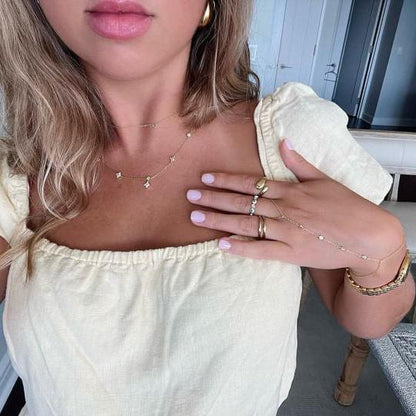 A woman wearing multiple pieces of delicate gold jewelry, including a gold hand chain with distanced cubic zirconia stones in bezel settings, draped elegantly across her hand and fingers. She is also wearing a layered gold necklace and several gold rings, creating a stylish and cohesive look. The woman is dressed in a soft yellow, off-shoulder top, complementing the warm tones of the gold jewelry, with the background showing a chic, modern interior setting.