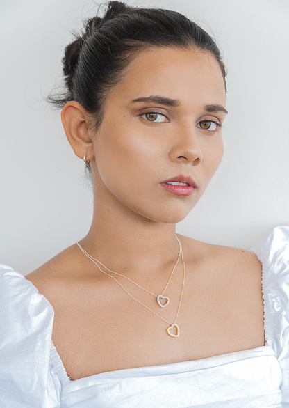 A model wearing a layered necklace featuring two heart charms, one gold and one silver, encrusted with stones. The necklace stands out against the model's simple white top, creating a polished and elegant look.