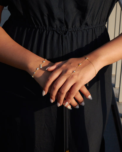 A model wearing the In Bloom Hand Chain and a matching bracelet, both crafted in gold-plated sterling silver with delicate cubic zirconia floral accents. Her hands are elegantly crossed, showcasing the dainty jewelry against a black dress, with soft sunlight highlighting the gold details.