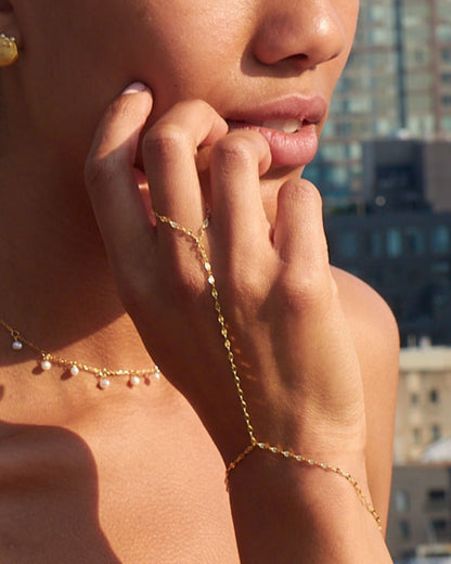 A model wearing the Feather Hand Chain in gold, with the chain elegantly tracing from the finger to the wrist. She poses against a city skyline, bathed in warm sunlight, highlighting the chain’s delicate yet eye-catching shimmer. She is dressed in a red strapless outfit, adding a bold contrast to the gold jewelry.