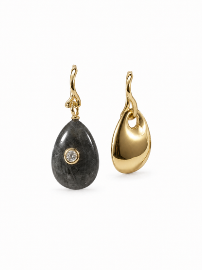 Mismatched yellow-gold wavy hoop earrings with removable charms: one full-width dark natural-stone teardrop with a clear bezel-set stone and one polished sculptural gold teardrop, on white.