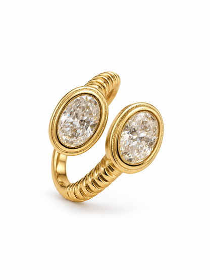 Front three-quarter view of an open yellow gold-tone ring with two oval moissanite stones in bezel settings and ribbed shoulders