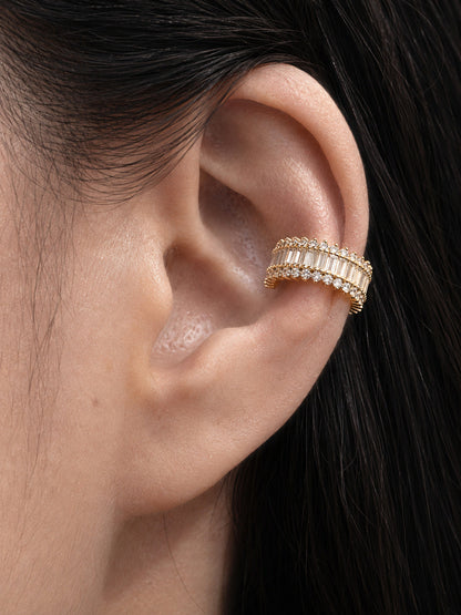 Realistic close-up of the Piercing Optional embellished ear cuff worn along the inner conch, showing its 18K yellow gold vermeil setting, central row of white baguette-cut stones, round stone borders, and piercing-free fit against naturally textured skin and dark hair.