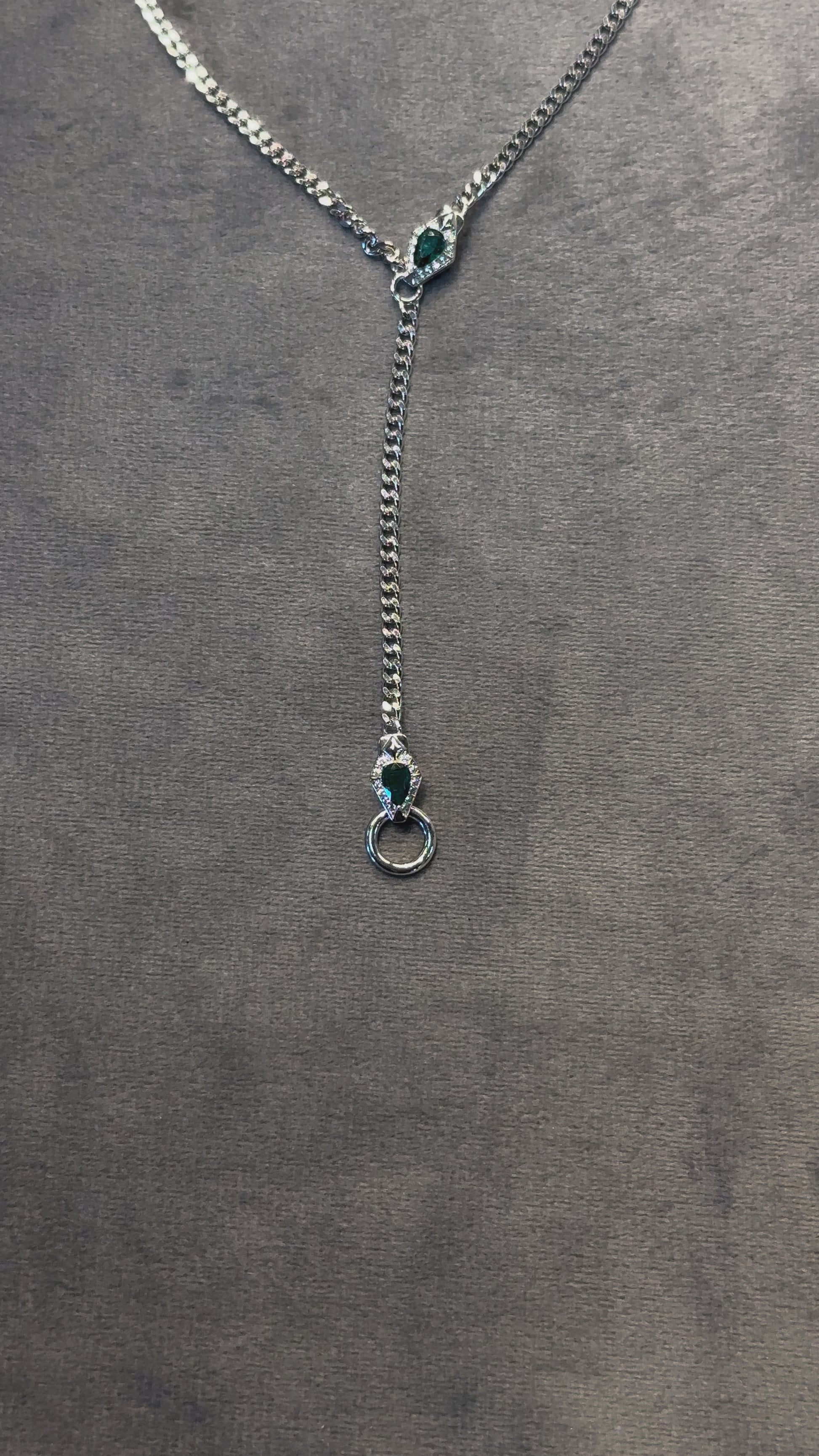 video of convertible/modular necklace featuring emerald green accent and dainty curb chain in platinum/ white gold finish, displayed in a lariat shape on a grey background. The video moves from bottom to the top to show details of the jewelry.